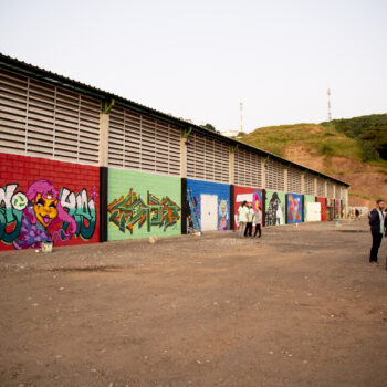 Foto: Alícia Peres | Descrição: Foto da lateral do longo galpão da Avemare, com mais de dez painéis grafitados no muro. À frente do galpão, um pátio de areia com algumas pessoas circulando. Ao fundo, um morro com vegetação e o céu cinza.