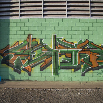 Artista: Morg | Descrição: Grafite na parede esverdeada da lateral do galpão de um grafismo em tons de verde escuro e laranja. A tipografia tem linhas retas, contornadas de branco, com efeito 3D. As letras formam a assinatura “Peres”, de maneira estilizada, com as letras em verde, levemente na horizontal, sombreadas em laranja. No canto inferior direito, a assinatura.