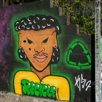 Artista: KS | Foto: Rogério Vieira e Léu Britto. Descrição - Sobre muro preto, grafite de um jovem afro-indígena, visto do peito para cima. Ele tem cabelo cacheado preto, raspado nas laterais e longo à altura da nuca; rosto ovalado, olhos puxados pretos, nariz largo, boca carnuda. Usa uma camisa amarela com a palavra “Recicle”, em verde. À direita, o símbolo da reciclagem, em verde, e, abaixo, a assinatura em branco.