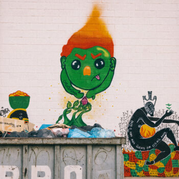 Artistas: Ayô, Nabrisa e Nzinga | Foto: Bruno Jungmann | Sobre uma parede branca, três grafites de estilo diferentes. Eles são vistos parcialmente. A frente, um container de ferro cheio de reciclados. Da esquerda para direita, do primeiro grafite vê-se a testa de uma mulher com um balde na cabeça, cheio de frutas. À direita, um ser verde com cabelo laranja, esfumado no alto em forma de cone. Ele tem olhos redondos pretos, sobrancelhas laranja e nariz laranja mais claro. Está com a língua presa do lado da boca e com os braços para frente em direção a um botão de rosa que brota da terra. O terceiro grafite é de uma silhueta de uma mulher nua sentada sobre um chão de terra em gomos verde, vermelho e amarelo. A mulher tem no colo uma esfera amarela em chamas. Sobre o corpo negro está escrito em branco, das costas até os joelhos, "2023 o ano mais quente em 125 mil anos". O rosto dela é coberto com desenhos tribais brancos e tem um arranjo na cabeça em forma de coroa.